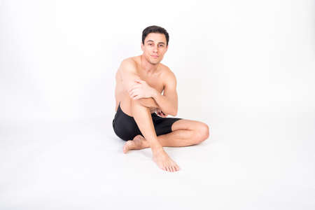 Shirtless athlete man sitting on the floor looking at camera. Full body, white background.