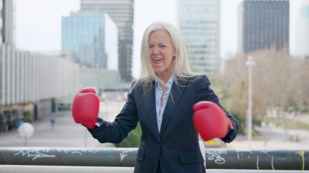 Mature aggressive businesswoman screaming clashing boxing gloves outdoorsの写真素材