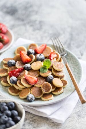 Tiny pancakes for breakfast. Cereal pancakes with blueberries, strawberries in plate on grey background. Trendy food. Selective focusの写真素材