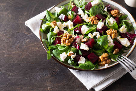 Beet or beetroot salad with fresh arugula, soft cheese and walnuts on plate, dressing and spices on dark wooden background, copy space, top viewの写真素材