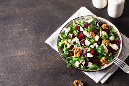 Beet or beetroot salad with fresh arugula, soft cheese and walnuts on plate, dressing and spices on dark wooden background, copy space, top viewの写真素材