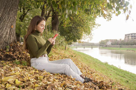 Happy smiling woman using phone on the autumn embankment near the riverの写真素材