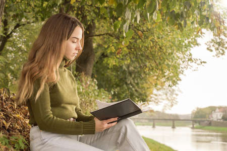 peaceful woman reading book. autumn season. female with trendy clothes relaxing near water and reading book. girl enjoying the weekend.の写真素材