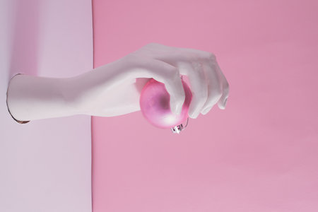 Creative arrangement with artificial woman's hand reaching out of a white wall holding pink Christmas decoration. Pastel background. New Year evening composition.の写真素材