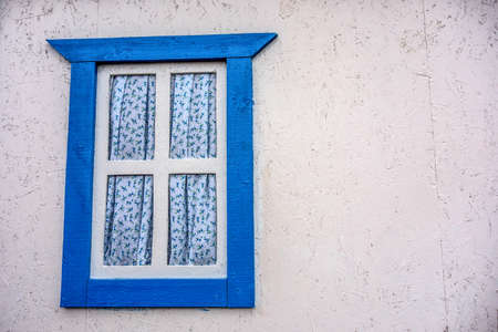 Blue wooden window of a Ukrainian hut with floral curtains close-up against a white wall.