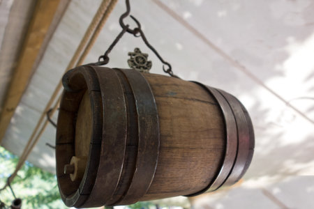 Old wine barrel with iron rings is suspended from a metal hook under a white canopy of a tent. A beautiful background for advertising oil, wine and strong alcoholic drinks. copyspace.の写真素材