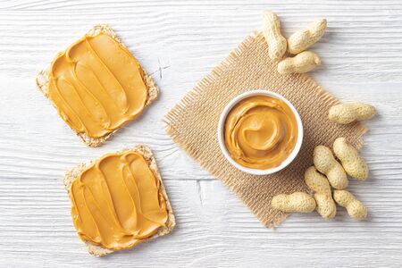 Creamy peanut butter. Paste in bowl, sandwich and peanuts on white wooden background. Above view.