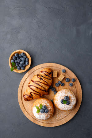 Breakfast - coffee, croissant, sweet donuts (buns) with powdered sugar and blueberries. Dark gray background. Flat lay.の写真素材