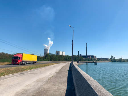 Red and yellow truck in motion on a road next to thermal power plant dam with coal facilities and steaming cooling tower on backgroundの写真素材