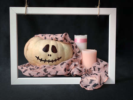 Adorable white Halloween pumpkin with a pink scarf next to white and pink candles in white frame on black backgroundの写真素材