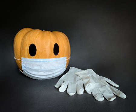 Halloween and the Coronavirus. Orange Halloween pumpkin wearing medical mask next to rubber gloves on dark gray backgroundの写真素材