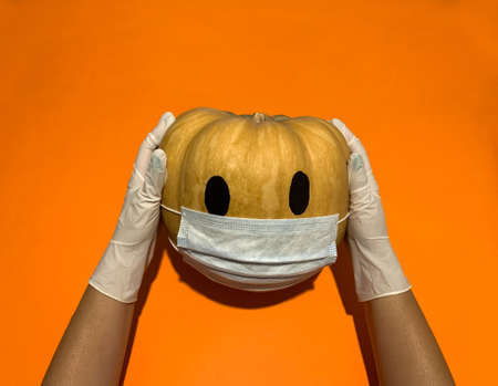 Coronavirus protection in Halloween 2020. Two hands wearing rubber glove holding pumpkin with medical face mask over orange backgroundの写真素材