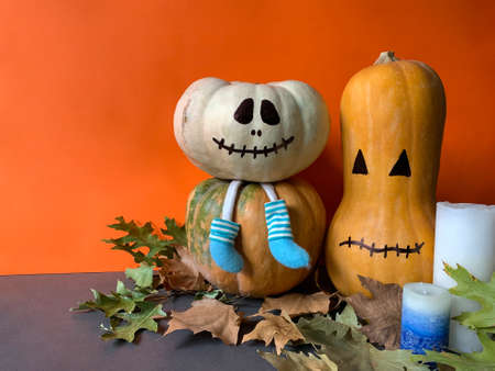 Cute Halloween wallpaper with white pumpkin with legs wearing blue socks, orange long pumpkin and candles against orange backgroundの写真素材