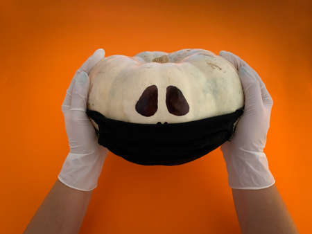 Halloween 2020 with Coronavirus. Hands with medical gloves holding white pumpkin with black protective mask on it against orange backgroundの写真素材