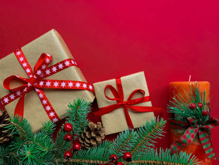 Christmas decorations layout with gifts wrapped in craft paper with red ribbons, fir branches, cones, berries and holiday candle on red background.の写真素材