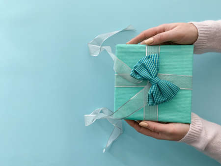 Close up hands giving blue present with big bow. Top view of gift box over pastel blue background with copy spaceの写真素材
