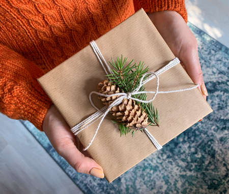 Hands of woman holding christmas gift box wrapped in kraft paper, decorated with fir tree branch and pine cone.の写真素材