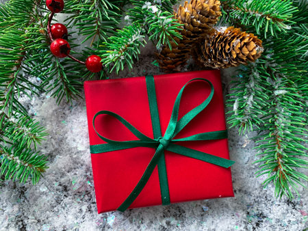Top view New year or Christmas gift box with red paper and green gibbon. Holiday frame with green fir tree branches, pine cones and red berries on snow.の写真素材