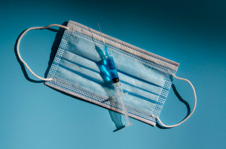 Syringe with coronavirus vaccine in medicine mask on blue background, medicine and drugの写真素材