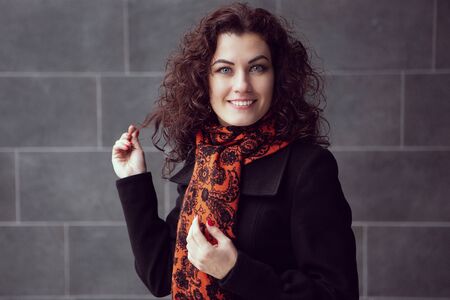 A beautiful smiling girl looks at the camera touching her hair. Portrait of a woman in a dark coat and a bright scarf. Romantic woman Beautiful gentle model.の写真素材