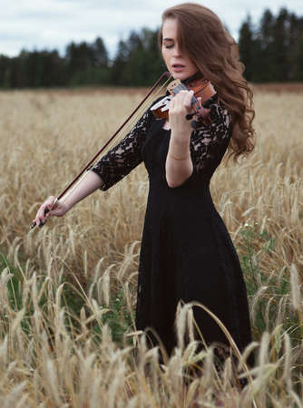 sexy young woman in a black dress enthusiastically playing the violin in a wheat field in a strong windの写真素材