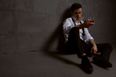 Portrait of a thoughtful handsome young man with a glass of alcohol in his hands sitting on the floor against a stone wall. The concept of pastime, rest and relaxationの写真素材