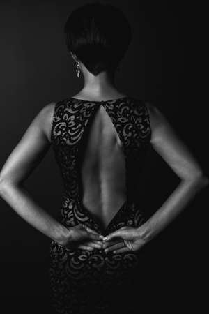 A sexy woman is standing with her back to the camera, undressing or zipping up her black tight dress, photo on a black background. Fashion, sexy stylish image for women conceptの写真素材