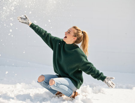 Young woman in casual winter clothing squatting in deep snow, joyfully throwing fresh white snow into the air during a sunny day in an outdoor winter landscapeの素材