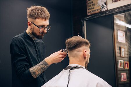 
Making haircut look perfect. Young bearded man getting haircut by hairdresser while sitting in chair at barbershop. tattoo.の写真素材