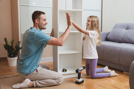 father and daughter give a cheerful high five while assembling a white shelf together on the floor, surrounded by tools and in a bright modern living roomの写真素材