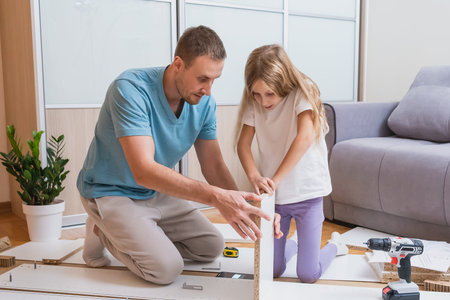 father watches as his daughter intently connects two large furniture panels on the floor surrounded by tools, parts, and instructions in a modern living roomの写真素材