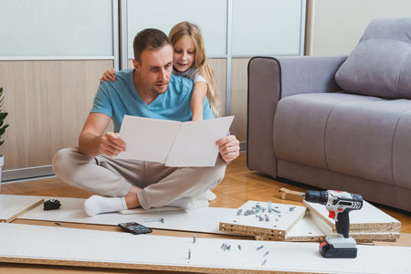 father studies assembly instructions with a puzzled look as his daughter hugs his shoulders and...