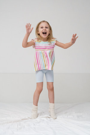 A smiling girl jumps on a bed with arms spread out in a bright minimalist room wearing a colorful rainbow striped shirt joyful energetic childhood play home family lifestyle conceptの写真素材