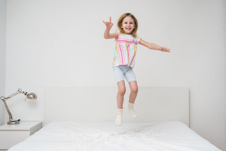 A smiling girl jumps on a bed with arms spread out in a bright minimalist room wearing a colorful rainbow striped shirt joyful energetic childhood play home family lifestyle conceptの写真素材