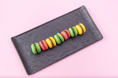 Top View of Tasty french macarons in a black serving plate on pink pastel background. Red, yellow and green macaronsの写真素材
