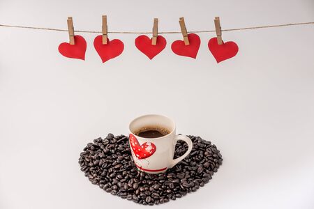 Cup of Coffee with heart made of coffee grains on white background and Clothes pegs and red paper hearts on rope above. Valentines day conceptの写真素材