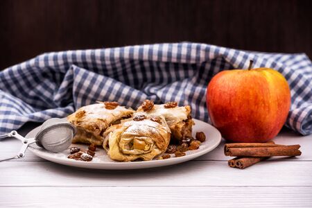 Slices of homemade Apple pie strudel in a plate with ingredients on a rustic white wooden table. Pie with apples, raisins and cinnamon, sweet pastriesの写真素材
