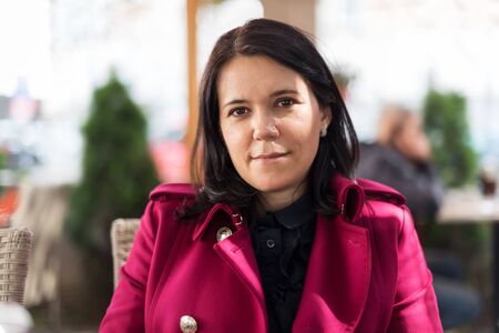 Image of smiling beautiful adult woman in wearing a pink coat sitting in street cafe restaurant outdoorsの写真素材
