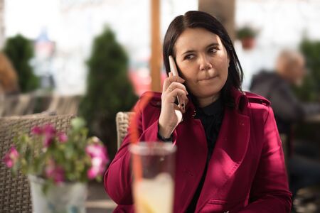 Image of a beautiful adult woman wearing a pink coat talking on a cellphone while sitting in street cafe restaurant outdoorsの写真素材
