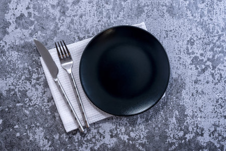Black plate, cutlery with utensils and napkin on rustic stone table top view. Table setting.の写真素材