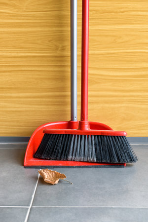 Red handled broom with red dustpan for cleaning stand near the wall with a single yellow leaf in front. Close up.の写真素材