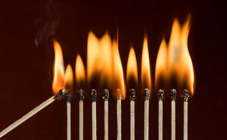 Lit match next to a row of lighting matches. Red phosphorus matches on dark red background. Concept of ignition or initiationの写真素材