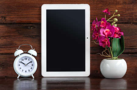 Tablet with Alarm clock and artificial flower decoration on a dark wooden shelf. Clipping path for tablet screenの写真素材