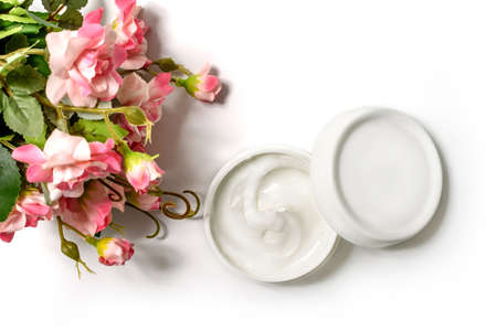 White Jar and white cap with body cream on white background with artificial rose flowers. White jar with face cream isolated. Cosmetic product for skin care. Top Viewの写真素材