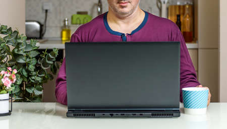 Trendy Mature man working from home with laptop sitting at the table in his kitchen, dressed in Purple Pajamas. Home office conceptsの写真素材