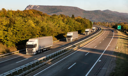 Convoy or caravan of transportation trucks passing on a highway at sunset. Highway transit transportation with red and yellow lorry trucksの写真素材