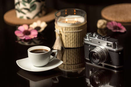 Coffee scene with vintage camera. A cup of coffee and vintage photo camera on a dark glass table with 

candles and flowers. Still life concept with coffee and old cameraの写真素材