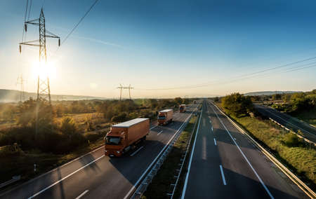 Sunset Highway transit. Convoy or caravan of Orange transportation trucks passing on a highway at amazing sunset. Highway transportation with lorry trucksの写真素材