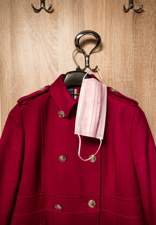 Red womans coat and Protective surgical face mask on anteroom coat hanger stand during pandemicsの写真素材