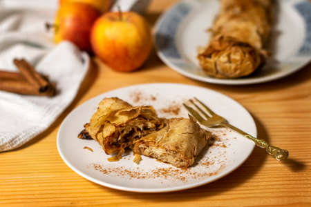A roll of homemade baked fresh apple pie strudel with an appetizing slice of apple pie on the plate and fresh apples and cinnamon sticks on a wooden table. Rustic breakfast conceptの写真素材